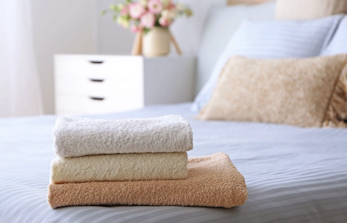 Three neatly folded towels in beige, cream, and white are stacked on a bed with a blurred background showing a nightstand and flowers.