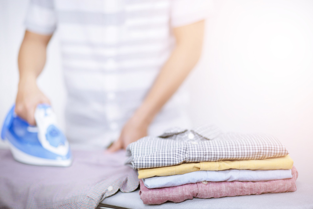 A person ironing a shirt with a blue iron, with a neatly folded stack of shirts in the foreground.