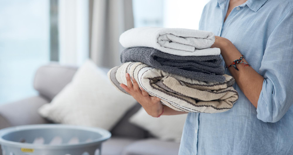 A person in a blue shirt holds a stack of folded towels, with a laundry basket and sofa visible in the background.