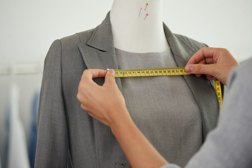 A person measures the chest of a dressmaker's mannequin dressed in a grey blazer using a yellow measuring tape.