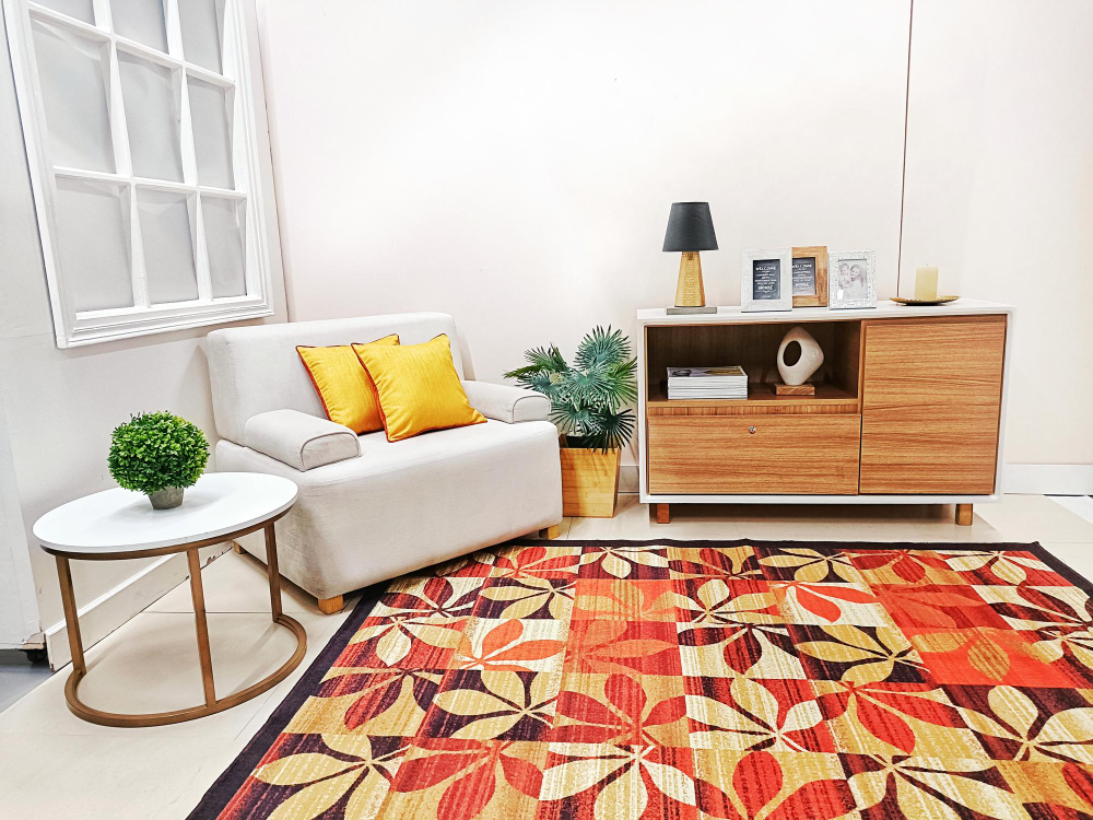 A modern living room with a beige armchair, yellow cushions, a white side table with a plant, a patterned rug, a wooden cabinet, and decorative items against a white wall.