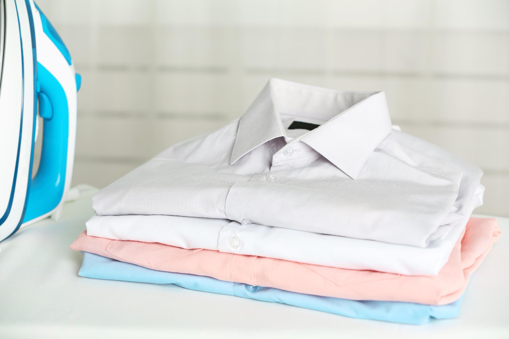 A neatly folded stack of shirts in white, pink, and blue colors sits beside a blue and white iron on a white surface.