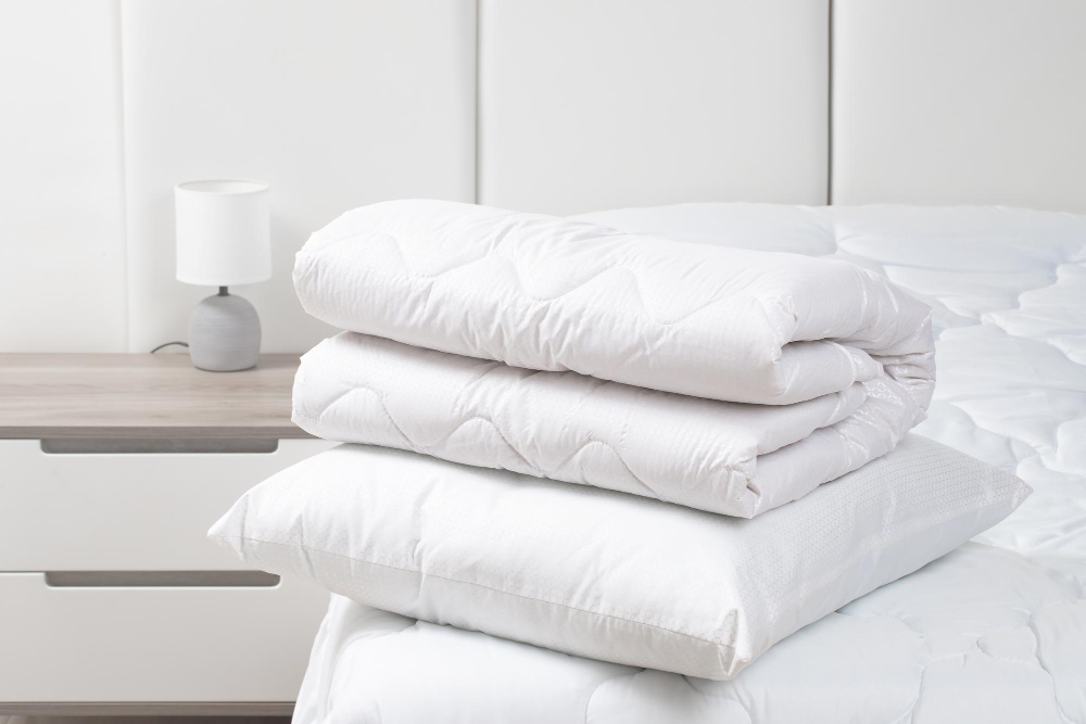 A folded white comforter stacked on a white pillow, placed on a bed next to a nightstand with a lamp.