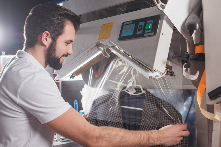 Man operating a dry cleaning machine, covering a plaid suit jacket with plastic wrap in a laundry facility.