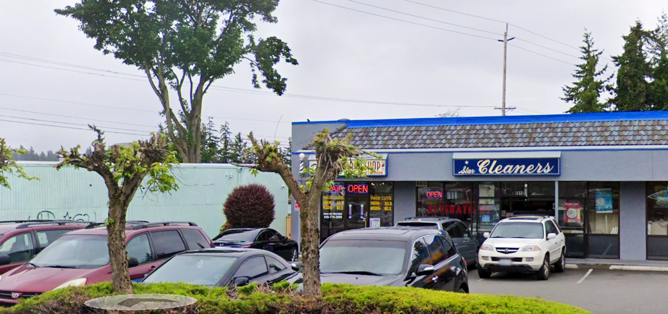 A small shopping plaza parking lot with several parked cars in front of a laundromat and cleaners storefront on a cloudy day.