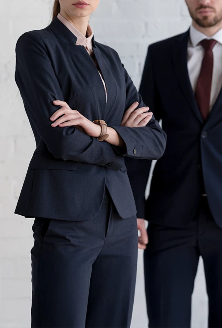 Two people in business suits standing indoors, one with arms crossed in the foreground and the other standing slightly behind. Their faces are partially out of frame.