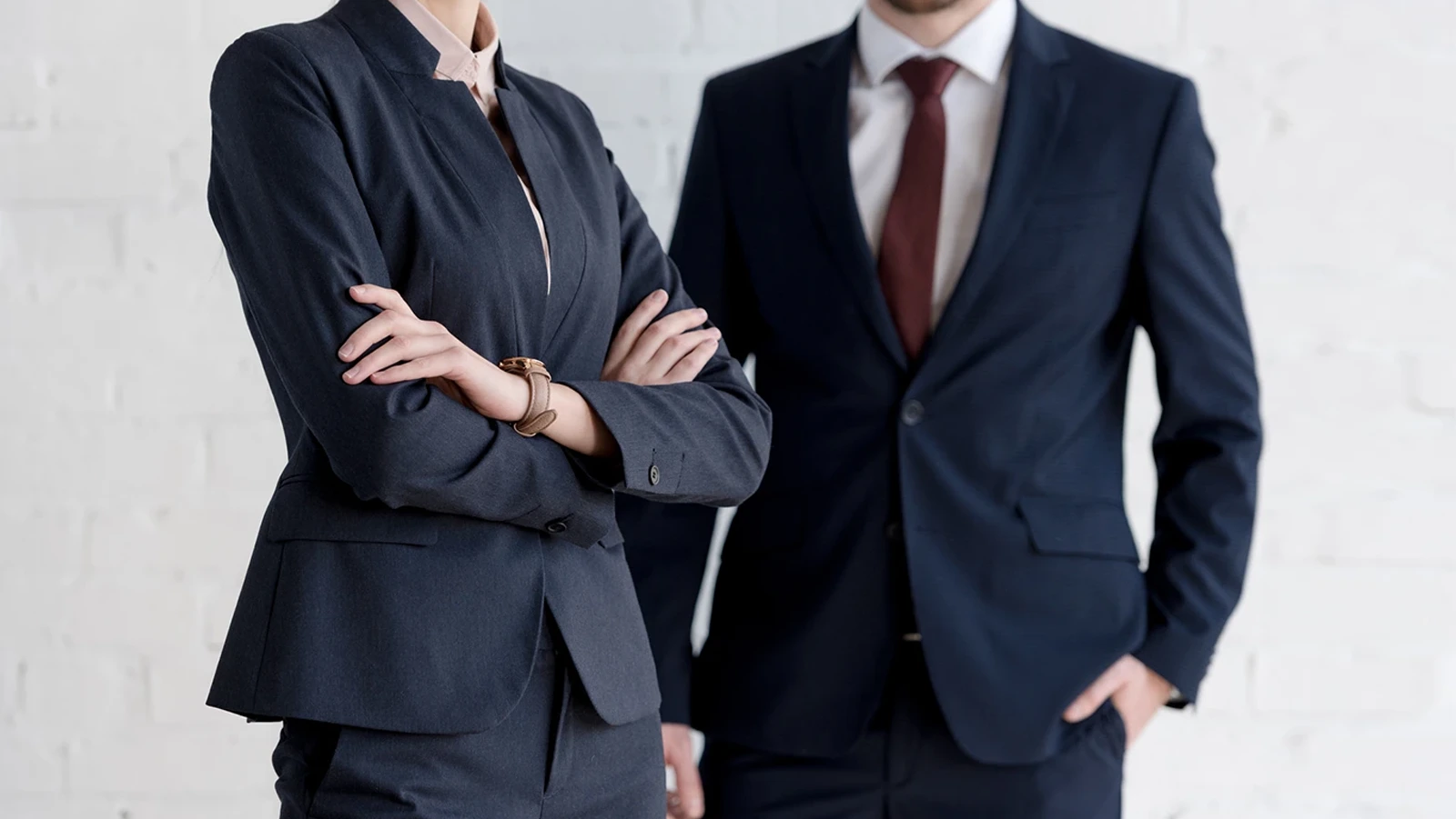 Two people in business attire, one with arms crossed and the other with hands in pockets, stand against a white brick wall. Both wear dark suits and light shirts with ties.