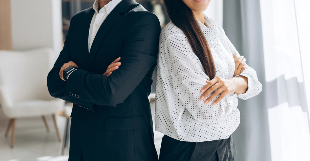 Two people standing side by side with arms crossed, dressed in business attire, partially facing the camera in a bright office setting.